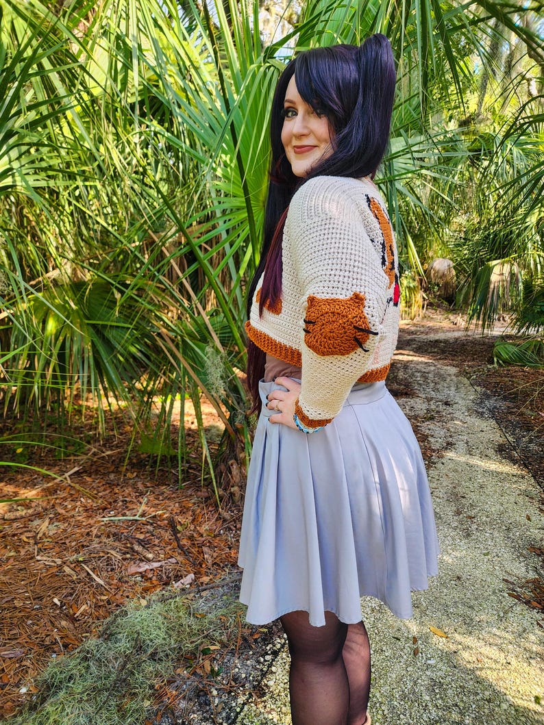 May include: A woman wearing a white and brown crocheted cropped sweater with a cat design. The sweater has a brown cat paw on the sleeve. She is wearing a light blue skirt and black tights.