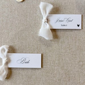 May include: Two rectangular white paper place cards with frayed-edge white fabric bows attached. One card reads "Jaime Coast Table 1" with a small chicken icon, and the other reads "Bride" in black script. Several small round wax seals are scattered nearby.