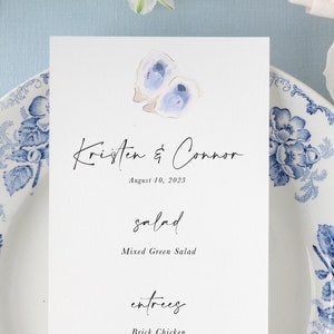 May include: A wedding menu card with a watercolor illustration of oysters and a lemon. The menu includes salad, entrees, sides, and dessert. The text reads "Kristen & Connor" and "August 10, 2023".