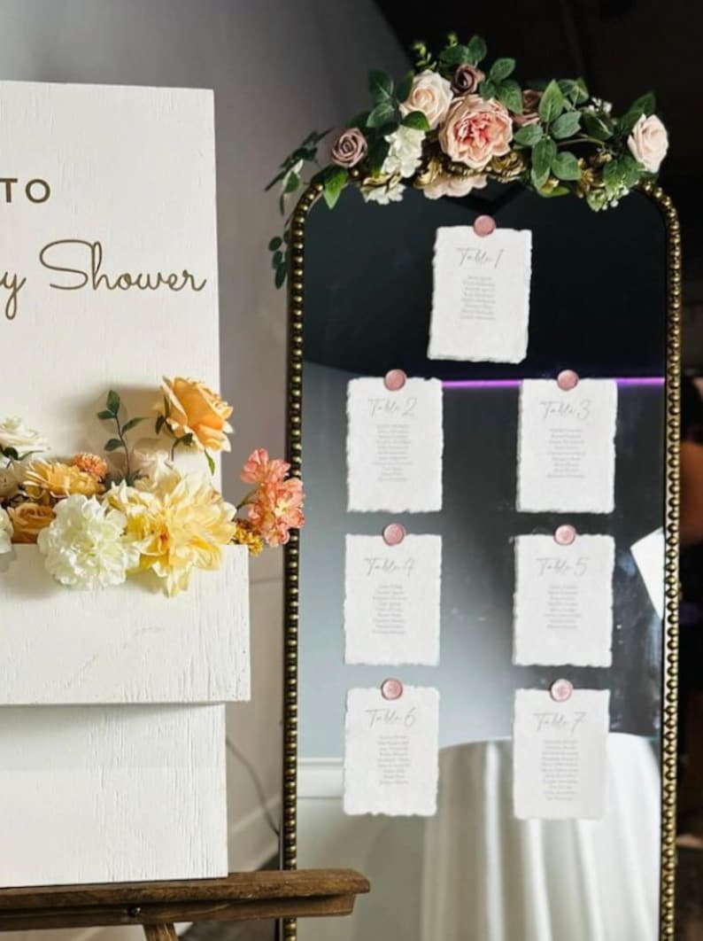 May include: A gold mirror with a beaded frame decorated with pink and white flowers. Seven white cards with handwritten table numbers are attached to the mirror with pink wax seals.