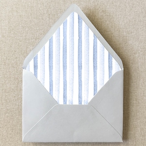 May include: A light gray envelope with a watercolor-style interior lining featuring vertical blue stripes on a white background. The envelope is open, revealing the striped pattern. The envelope is on a neutral-toned surface.