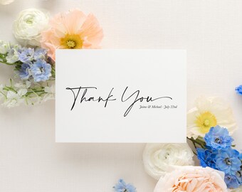 May include: White card with black calligraphy that says "Thank You" and the names "Jaime & Michael" and the date "July 22nd". The card is surrounded by white, peach, and blue flowers.