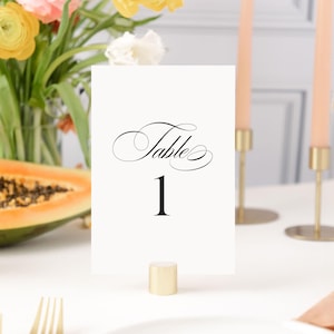 May include: White card with black text that reads "Table 1" in a cursive font. The card is on a gold stand.