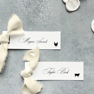 May include: Two white place cards with black calligraphy names and animal silhouettes. The top card reads "Megan Sorrels" with a chicken silhouette. The bottom card reads "Taylor Beck" with a cow silhouette.