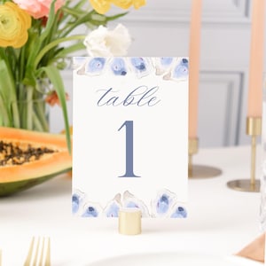 May include: A white table number card with the word "table" in script and the number "1" in a soft blue hue. The card is decorated with watercolor blue floral accents and sits in a gold-colored holder. Flowers and candles are in the background.