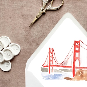 May include: An overhead shot of stationery items. A white envelope with a watercolor illustration of the Golden Gate Bridge is open. Gold scissors and white wax seals with initials are scattered around the envelope on a mauve surface.