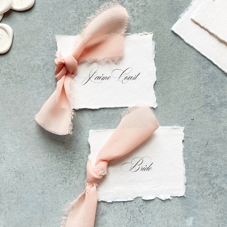 May include: Two white cards with torn edges, each tied with a pale pink ribbon. The top card reads "Jaime Coast" and the bottom card reads "Bride".