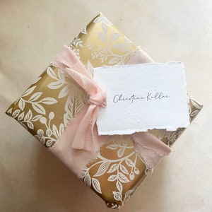 May include: A gold gift box with a white floral pattern is wrapped with a pink ribbon and a white tag that says "Christine Kollar".