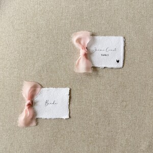 May include: Two rectangular white paper place cards with deckled edges, each adorned with a soft pink silk ribbon tied in a bow. One card features the text "Jaime Coast", "Table 1", and a small black chicken icon. The other card displays the text "Bride".