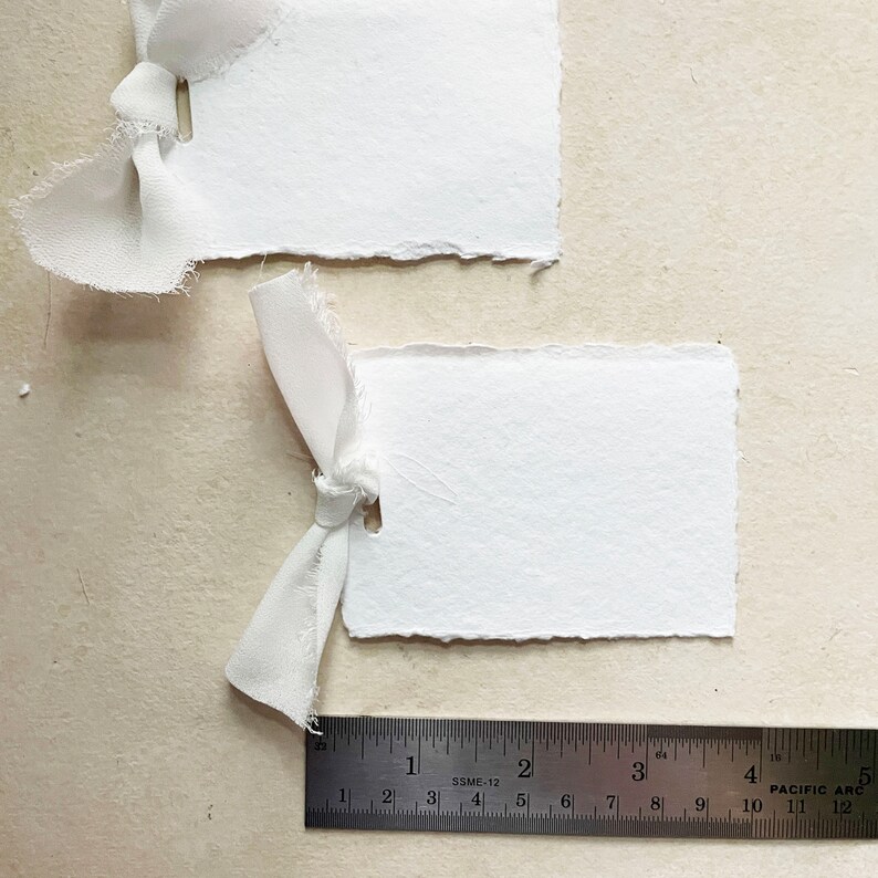 May include: Two white paper tags with frayed edges, each tied with a white ribbon. A ruler is placed below the tags, showing measurements in inches.