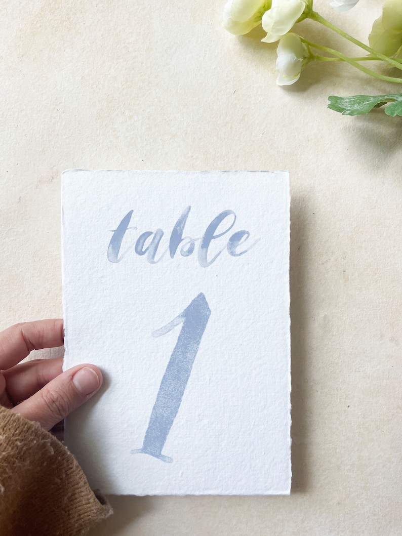 May include: A white card with the word "table" written in blue script and the number "1" written in blue watercolor.
