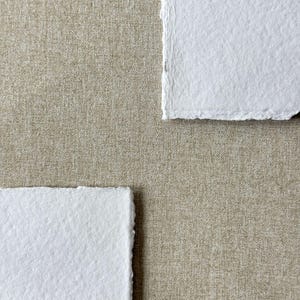 May include: Two rectangular sheets of bright white paper with rough, deckled edges are positioned diagonally against a solid, neutral-toned beige fabric background that features a visible, fine-woven linen-like texture throughout the surface.