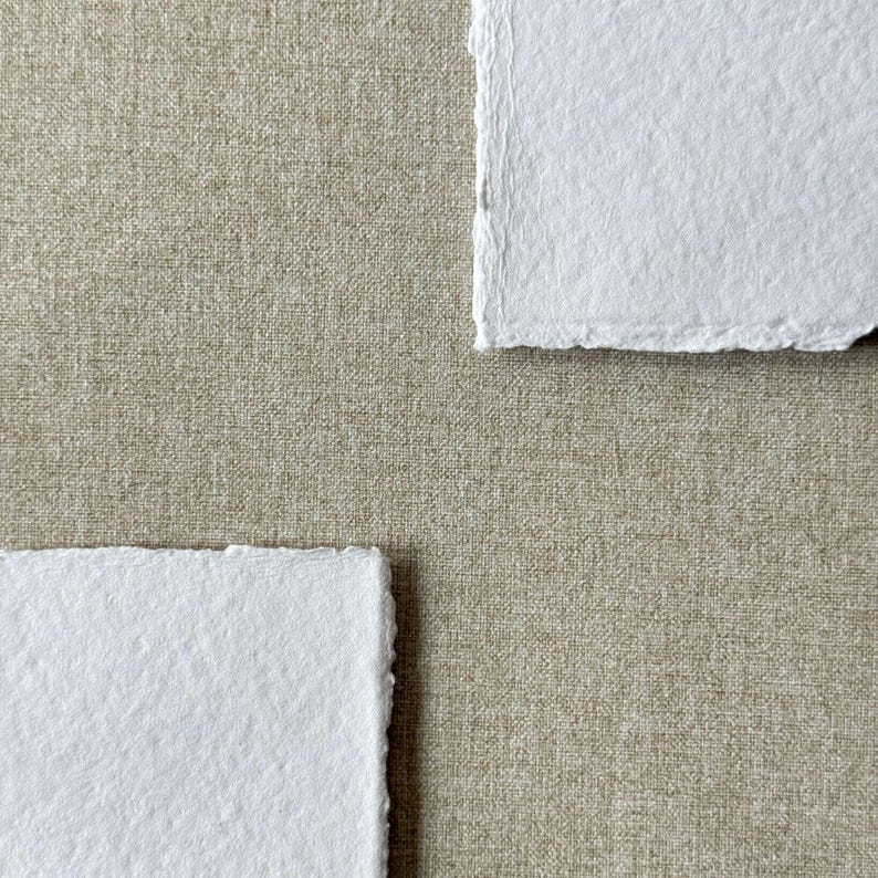 May include: Two rectangular sheets of bright white paper with rough, deckled edges are positioned diagonally against a solid, neutral-toned beige fabric background that features a visible, fine-woven linen-like texture throughout the surface.
