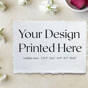 May include: A white card with the text "Your Design Printed Here" and the text "multiple sizes - 3.5x5" 4x6" 4x9" 5x7" 8x10" printed on it. The card is sitting on a light beige surface with white flowers scattered around it.