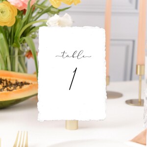 May include: White table number card with the text "table 1" in black script font. The card is on a gold stand and is sitting on a white table.