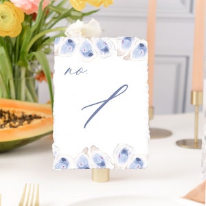 May include: A white table number card with a blue floral border and the text "no. 1" written in blue ink. The card is on a gold stand and is on a white table.