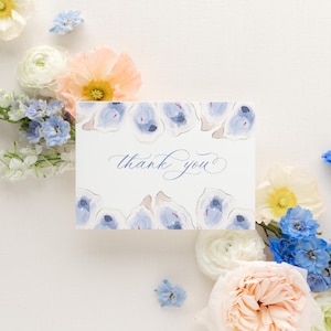 May include: A white thank you card with a blue and white watercolor design of oyster shells. The card is surrounded by a variety of flowers, including peach, white, and blue.