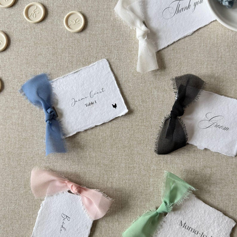 May include: Four rectangular white paper place cards with deckled edges, each featuring a coloured chiffon ribbon tied on the left side. The cards display printed text including names, titles, and a small black chicken icon, with matching buttons.
