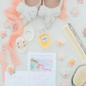 May include: Wedding details laid out on a white surface with a peach colored fabric. The details include a pair of white shoes with pearl bows, a perfume bottle, a comb, a hand mirror, a ring box, a seashell, and two wedding invitations. The invitations are for a wedding ceremony and reception. The invitations include the couple's names, the date, the time, and the location of the wedding. The invitations also include a RSVP card.