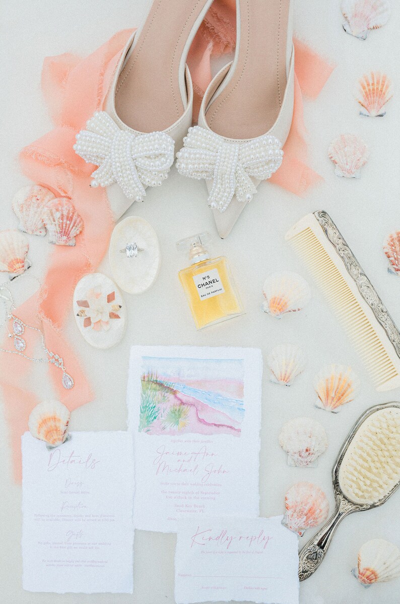 May include: Wedding details laid out on a white surface with a peach colored fabric. The details include a pair of white shoes with pearl bows, a perfume bottle, a comb, a hand mirror, a ring box, a seashell, and two wedding invitations. The invitations are for a wedding ceremony and reception. The invitations include the couple's names, the date, the time, and the location of the wedding. The invitations also include a RSVP card.
