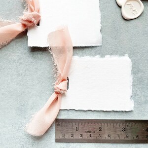 May include: Two white, textured, rectangular cards with scalloped edges. A pink ribbon is tied around one of the cards. A ruler is in the bottom of the image with the numbers 1, 2, 3, and 4 visible.