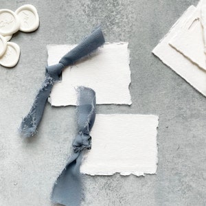May include: Two blank white cards with torn edges, tied with blue ribbon, on a gray textured background.