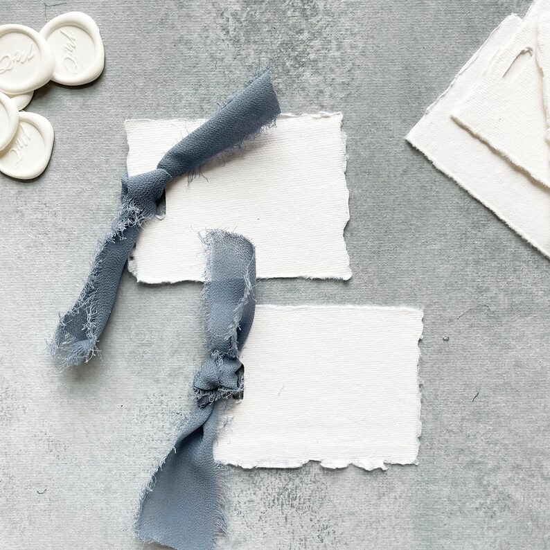 May include: Two blank white cards with torn edges, tied with blue ribbon, on a gray textured background.