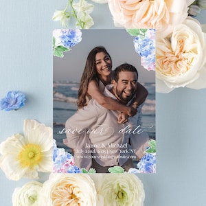 May include: Save the date card with a photo of a couple on a beach. The card is decorated with blue and pink watercolor hydrangeas. The text on the card reads "Save our date", "Jaime & Michael", "July 22nd, 2025 | New York, NY", and "www.yourweddingwebsite.com".