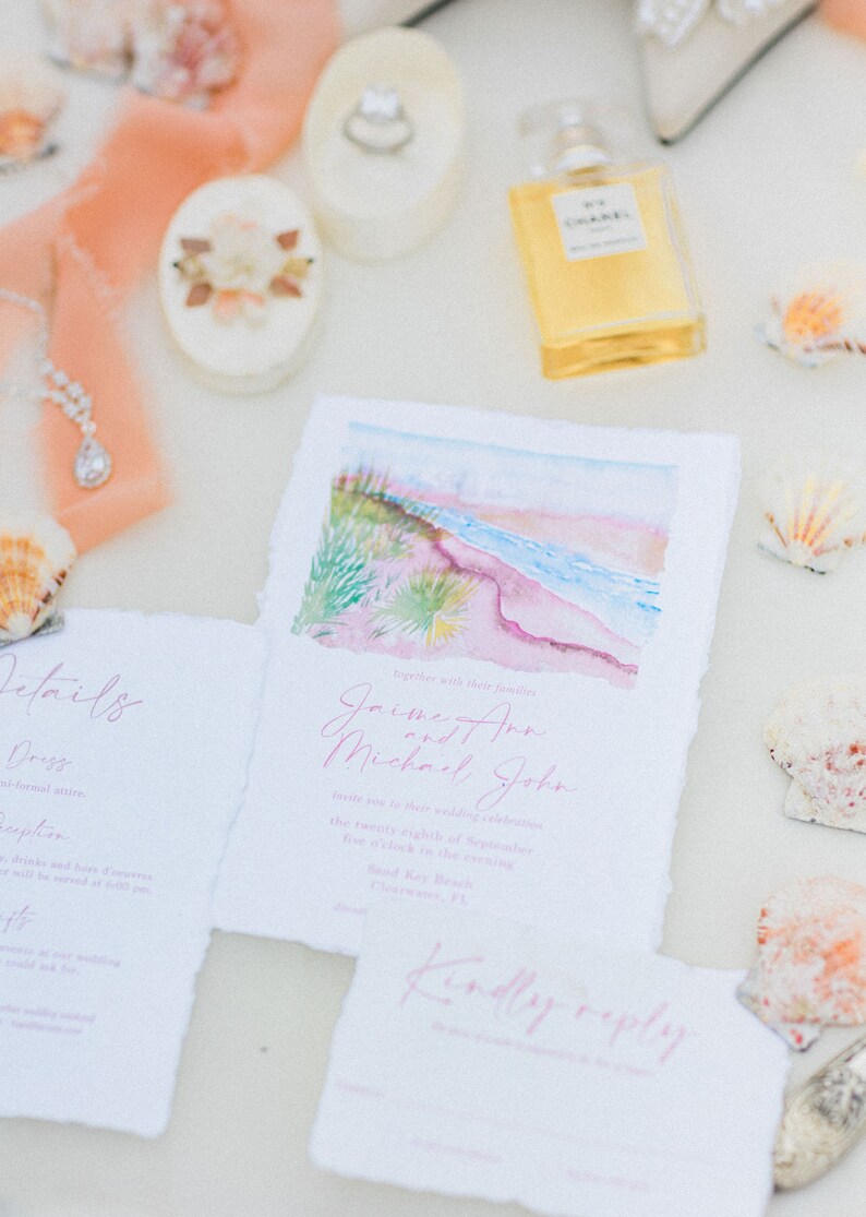 May include: A wedding invitation suite with a watercolor illustration of a beach scene. The invitation is printed on textured paper with pink and white text. The text reads "Together with their families,  Joime Ann and Michael John invite you to their wedding celebration the twenty-eighth of September five o'clock in the evening South Key Beach, Clearwater, FL. Kindly reply."