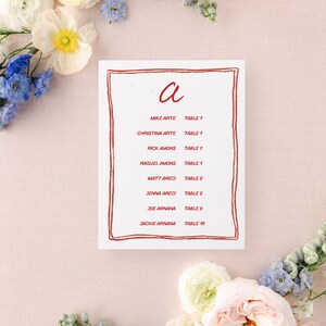 May include: A white card with a red border and the letter "a" in red script. The card lists names and table numbers for a wedding reception. The names include Mike Arte, Christina Arte, Rick Amons, Raquel Amons, Matt Areci, Jenna Areci, Joe Arnana, and Jackie Arnana.