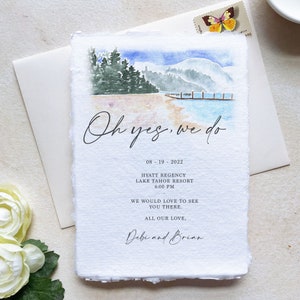 May include: Watercolor wedding invitation featuring a lake scene with mountains and a dock. The text "Oh yes, we do" is written in elegant script. Additional text includes the date, location, and names Debi and Brian.