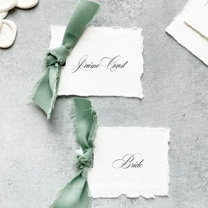 May include: Two white cards with torn edges, each tied with a sage green ribbon. The top card reads "Jaime Coast" in cursive script. The bottom card reads "Bride" in cursive script.