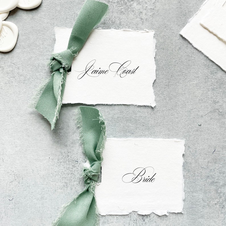 May include: Two white cards with torn edges, each tied with a sage green ribbon. The top card reads "Jaime Coast" in cursive script. The bottom card reads "Bride" in cursive script.