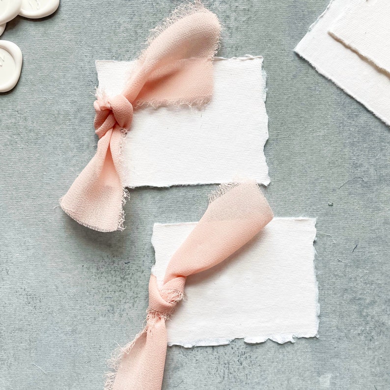 May include: Two white, textured, rectangular cards with frayed edges, each tied with a pale pink ribbon bow. The ribbons are made of a soft, sheer fabric.