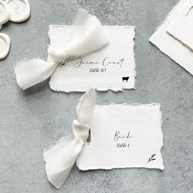 May include: Two white place cards with black calligraphy. The top card reads "Jaime Coast Table 10" with a small cow icon. The bottom card reads "Bride Table 1", with a small branch icon. Both cards are tied with white ribbon bows.