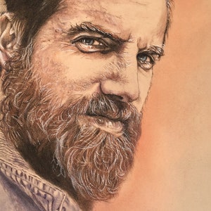 May include: A close-up portrait of a man with a brown beard and a light brown background. He is wearing a blue denim jacket and has a serious expression on his face.