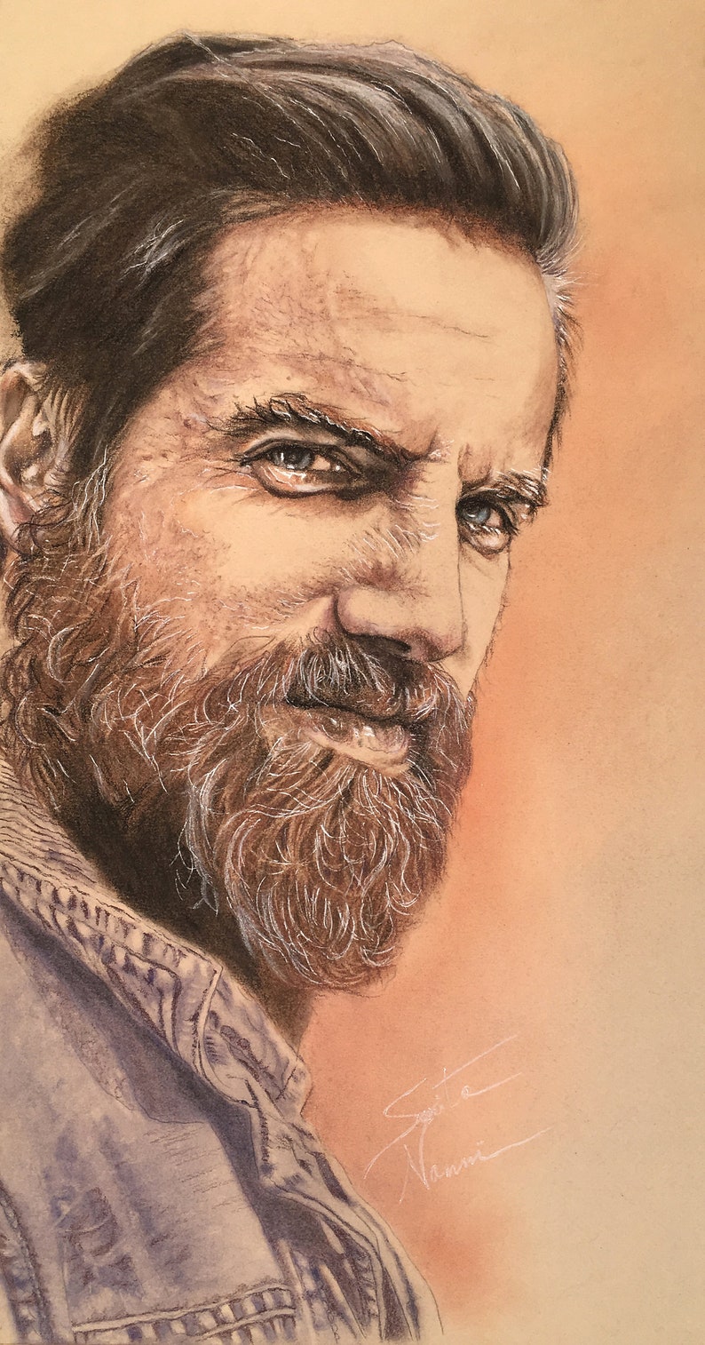 May include: A close-up portrait of a man with a brown beard and a light brown background. He is wearing a blue denim jacket and has a serious expression on his face.