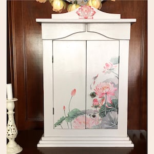 Könnte beinhalten: Ein weißer Schrank mit zwei Türen, verziert mit einem floralen Lotus-Design in Rosa und Grün. Der Schrank hat eine dekorative Oberseite mit einer Lotusblume und einen kleinen Sockel. Der Schrank ist weiß lackiert und hat eine Vintage-Ästhetik.