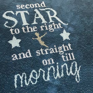Peut inclure: Broderie au point de croix sur tissu bleu foncé. Le texte dit "second STAR to the right and straight on till morning" en fil blanc. Une silhouette dorée d'une figure est au centre, avec deux formes d'étoiles.