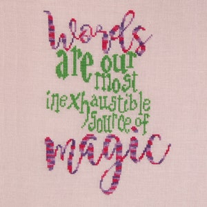 Op de afbeelding: Borduurwerk met kruissteek met de tekst "Words are our most inexhaustible source of magic" in groene en paarse draad op een roze stoffen achtergrond.