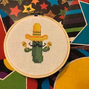 May include: A green cactus wearing a yellow sombrero and holding maracas. The cactus has a black mustache. The embroidery is framed in a wooden hoop.