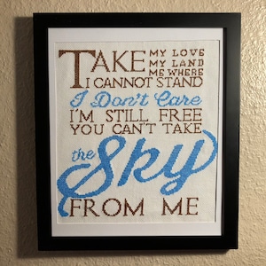 May include: Framed cross-stitch embroidery with the text "TAKE MY LOVE MY LAND ME WHERE I CANNOT STAND I Don't Care I'M STILL FREE YOU CAN'T TAKE the Sky FROM ME". The text is in brown and blue on a white background.