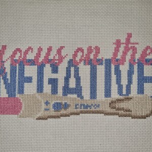 Op de afbeelding: Kruissteekkunstwerk op een lichtbeige stof. De tekst "focus on the NEGATIVE" is geborduurd in roze en blauw. Daaronder bevindt zich een beige zwangerschapstest met blauwe details en tekst.