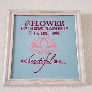 May include: A framed cross-stitch with a light blue background and pink flower design. The text reads "The flower that blooms in adversity is the most rare and beautiful of all."