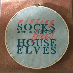 Puede incluir: Un bastidor de bordado de punto de cruz redondo, con la frase "Missing Socks Are a Sign of Freed House Elves" en coral y verde azulado. La labor está en un aro de madera.