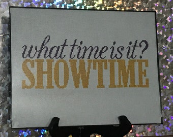 May include: A white framed sign with the text "what time is it? SHOWTIME" in purple and gold lettering.