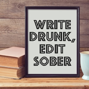 Puede incluir: Una impresión enmarcada con el texto "WRITE DRUNK, EDIT SOBER" en una fuente pixelada en blanco y negro. La impresión está sobre un fondo blanco y está enmarcada en un marco negro. La impresión está sentada sobre una mesa de madera con dos libros y una taza azul.