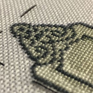 Puede incluir: Primer plano de un dise&ntilde;o de punto de cruz sobre tela blanca. El dise&ntilde;o presenta una secci&oacute;n de una forma clara delineada con hilo gris oscuro. La tela tiene una apariencia texturizada, con puntadas visibles que crean un patr&oacute;n detallado.