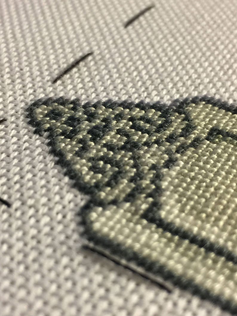 Puede incluir: Primer plano de un dise&ntilde;o de punto de cruz sobre tela blanca. El dise&ntilde;o presenta una secci&oacute;n de una forma clara delineada con hilo gris oscuro. La tela tiene una apariencia texturizada, con puntadas visibles que crean un patr&oacute;n detallado.