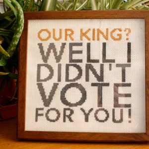 May include: Cross-stitch artwork in a wooden frame. The text reads "OUR KING? WELL I DIDN'T VOTE FOR YOU!" in orange, grey, and black thread on a cream background. The artwork is displayed on a wooden surface.